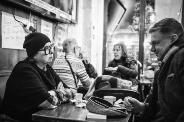 20221203-DSCF9348_MOD_BW_FRAME_resize Black and white photo of people sitting at a café table, engaged in conversation. A woman in glasses and a beanie faces a man wearing glasses—Fred & Adele enjoy their coffee as other patrons chat in the background.