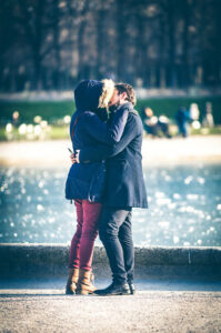 Remy and Barbara, wearing winter coats and boots, share a kiss by a body of water, with blurred trees and people in the background on a sunny day.
