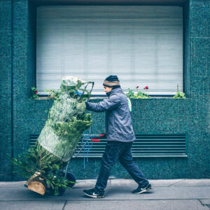 Marcel Tristan, wearing a beanie and mask, pushes a wrapped sapin on a hand truck along the sidewalk in front of a building with green tiles and a closed window.