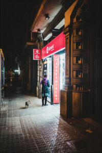A person stands at the brightly lit entrance of a supermarket at night, with a small chien on a leash beside them, while Marcus waits nearby on the quiet, dimly lit street.