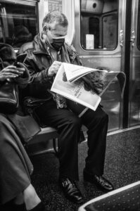 A man wearing a mask sits on a subway bench, reading a journal, reminiscent of scenes described by Pierre Delanoé. He is dressed in a jacket and dress shoes, while another person beside him uses a smartphone. The scene is in black and white.