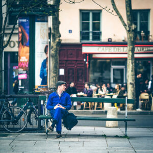Eric Maurice, dressed in a blue outfit and cap, sits alone on a green bench in a city square, looking to his left. Behind him are parked bicycles, a tree, and a busy cafe filled with people.