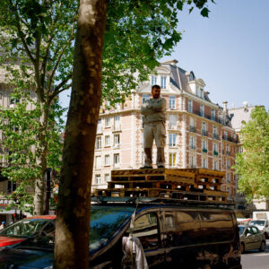 A man stands on stacked wooden pallets atop le toit de son camion noir parked on a city street, with apartment buildings and leafy trees in the background on a sunny day.