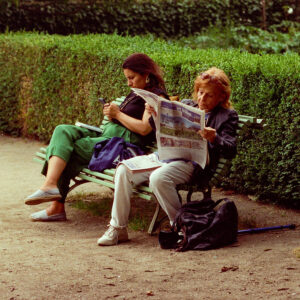 Cécile and Sonya sit on a park bench; Cécile reads a newspaper while Sonya looks at her phone. A green hedge stands behind them, with bags at their feet. The scene is set outdoors along a dirt path.