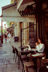 Viviane Chevalier sits at an outdoor café table on a city sidewalk, using a laptop. Other people walk along the street, while menu boards and awnings add to the relaxed, urban atmosphere.