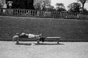 Mathias Marie lies on a bench in a park, resting with legs crossed and hands on their chest. Their shoes and a bag are on the ground nearby, with a grassy slope and stone balustrade in the background.