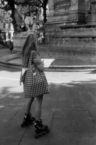 Melodie Perrin, in a checkered dress and rollerblades, stands by a fountain in a city square, sketching or writing on a clipboard. The black and white scene captures her creative moment.