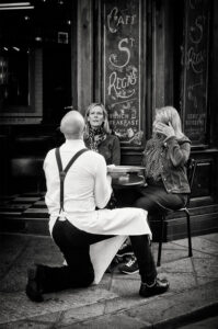 A garçon de café in a white apron kneels on the sidewalk, taking an order from two women seated at a small outdoor café table in front of a vintage-looking restaurant, creating a charming show for passersby.
