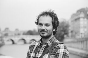 Daniel Latif, a man with curly hair and a beard, wearing a plaid shirt, stands outdoors near a river with a stone bridge and blurred buildings in the background. The photo is in black and white.