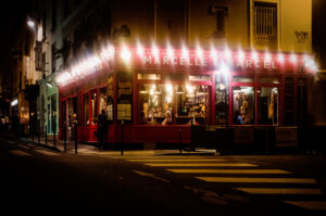A warmly lit restaurant called Marcelle et Marcel glows at night on a street corner, with diners visible inside through large windows and a few people strolling past outside.