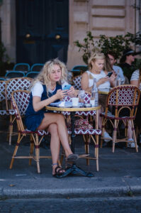 Two women sit at a sidewalk café, each looking at their phones. In the foreground, Cécile Rolland, with curly blonde hair and a white shirt under an overall dress, sits beside coffee cups and a water bottle on their table.