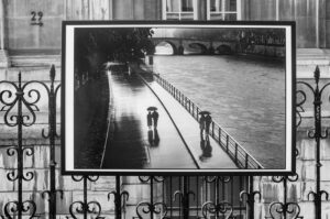 A black-and-white photo displayed on a wrought iron fence shows two couples with umbrellas walking along a riverside path in the rain, their reflections visible on the wet pavement, with a bridge in the background.