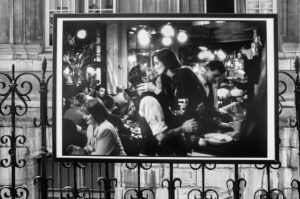 A black-and-white photograph displayed on an ornate metal fence, depicting a couple kissing passionately in a busy, dimly-lit restaurant or bar. Other patrons are seen in the background, engaged in conversation and dining.