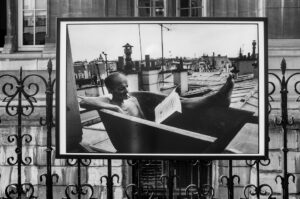 Black and white photo displayed outdoors, showing a man relaxing in a bathtub on a rooftop, reading a newspaper, with city buildings and antennas in the background. The framed photo hangs on a decorative metal fence.