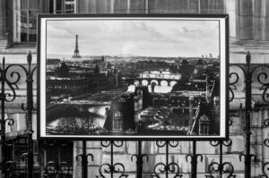 A black and white photograph is displayed on an ornate wrought iron fence. The image shows a scenic view of Paris with the Eiffel Tower in the distance and the Seine River flowing through the city, crossed by several bridges.