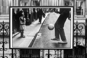 A black-and-white framed photo displayed on a balcony shows a young girl watching an adult play with a soccer ball on a street, with urban buildings in the background.