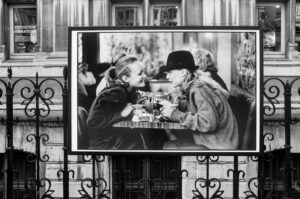 A black-and-white photograph displayed on an exterior fence shows two people sitting closely at a café table, engaging in conversation and smiling at each other. They each hold a cup, suggesting they are sharing a drink or coffee.