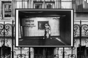 A black-and-white photo displayed on a metal fence shows a woman standing alone in an urban setting, with French signs on the wall behind her. The background includes a stone building with windows.