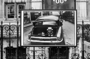 A black-and-white photo displayed outdoors shows three children smiling from the open trunk of a classic car, while another child stands beside it. The scene is framed by ornate metal fencing and historic buildings.