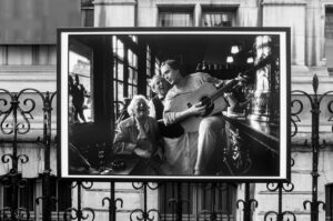 Black-and-white photo displayed outdoors shows an older woman laughing at a bar while a man beside her sings and plays guitar; another man stands between them, smiling. The display is mounted on a wrought-iron fence.