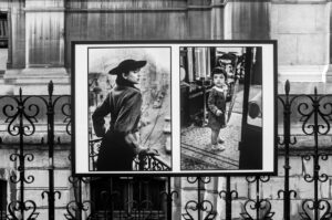 A black and white photo display mounted on an ornate metal fence, showing two vintage portraits: a woman in a hat looking sideways and a young child standing on a tram with a serious expression.