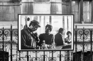 A black and white photo depicts an image of men socializing in what appears to be a café or bar. Two men in the foreground are engaged in conversation, while two others sit in the background. Bottles and glasses are visible on the table. The scene is framed by a fence.