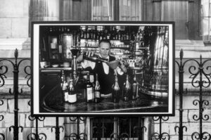 Black-and-white photo of a man behind a bar pouring liquid through a funnel into a wine bottle; the image is displayed on a fence in front of a building with large windows.