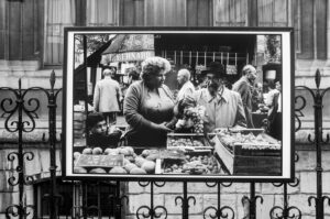 A black-and-white photograph displayed on a wrought iron fence shows a bustling market scene. An elderly man and a woman examine grapes at a fruit stall, while other market-goers browse in the background. A young boy stands next to them looking at the fruit.