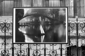 Black-and-white image of a photograph mounted on an ornate iron fence. The photograph depicts a sailboat on tranquil water with a bridge arching over in the background, surrounded by trees. There's a building visible behind the fence.