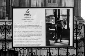 Black and white image of a display board in Paris with text titled "Une Lettre d'Amour à Paris" by Peter Turnley. A photograph shows a person standing, reading a newspaper. The display is outdoors, mounted on an ornate metal fence.