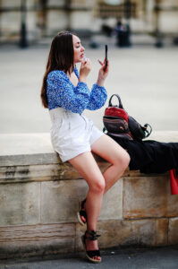 Daniella Aubry sits on a stone ledge in a blue floral blouse and white shorts, applying lipstick as she checks her reflection in a compact mirror. A black and red backpack and dark jacket rest beside her.