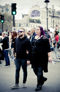 A couple walks arm in arm at an outdoor event; the man wears sunglasses, a black hoodie, and jeans, holding a drink, while the woman wears black clothes with purple hair and holds a can. People and buildings are in the background.