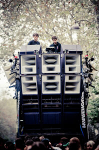 Two people stand behind a large wall of stacked speakers and audio equipment outdoors, elevated above a crowd, with trees and overcast sky in the background.