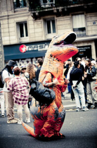A person in an inflatable dinosaur costume stands on a city street during a crowded event, holding a black bag, with people and buildings visible in the background.