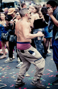 A man, shirtless with a large back tattoo and wearing sagging cargo pants revealing red underwear, dances in a lively street crowd. He holds a spray can and stands amid scattered colorful confetti, surrounded by people taking photos and enjoying the event.