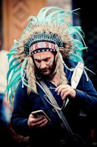 A man wearing a blue feathered headdress with intricate beadwork and face paint looks down at his smartphone. He is also dressed in a navy blue jacket and carrying a small bag across his shoulder.