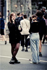 A young woman in black holds a drink and a cigarette, standing on a street beside another woman in light jeans. Several people walk away in the background, all wearing casual clothes.