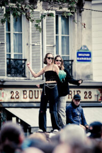 Two individuals stand on a structure amidst a crowd, dancing and smiling. One person is wearing a black crop top and jeans, while the other is in a dark jacket and sunglasses, holding a feathered green item. Behind them is a building with shuttered windows and a street sign.