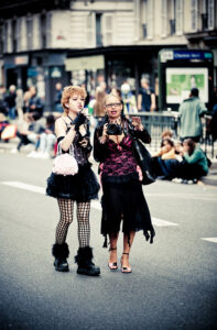 Two women in bold, eclectic outfits walk down a city street. One holds a camera, both wear black and pink clothing with unique accessories. Blurred crowd and buildings can be seen in the background.