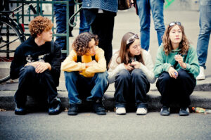 Four young people are sitting on a curb, engaged in conversation and looking relaxed. They are wearing casual clothing, with one holding a phone and another holding a drink. Bicycles and pedestrians are visible in the background.