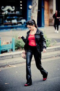 A woman in a black leather jacket and pants, red top, and red shoes walks on a city street, looking down. She carries a striped handbag and there are blurred storefronts and café lights in the background.