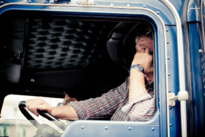 A person wearing a plaid shirt sits in the driver’s seat of a blue truck, resting their head on one hand and holding the steering wheel with the other, looking thoughtfully out the window.