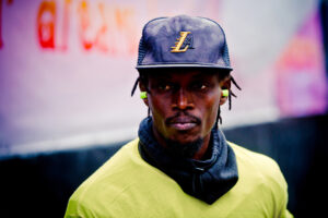 A man is wearing a black cap with an "LA" logo, neon green earbuds, a lime green shirt, and a black neck gaiter. He has a serious expression and the background is slightly out of focus with hints of mostly pink and red hues.