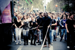 A diverse group of people dance and walk together down a city street during a parade or festival, with some holding long white poles at the front and colorful decorations in the background.
