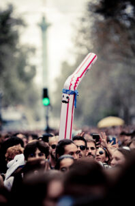 A crowd of people at an outdoor event. Notable among them is an inflatable tube man with a striped design, wearing sunglasses and a mustache, sticking up above the heads of the crowd. In the background, a green light can be seen, and trees line the street.