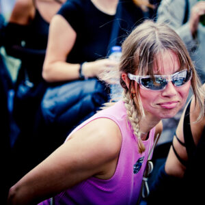 A woman with blonde hair in two braids wearing reflective sunglasses and a sleeveless purple top stands in a crowd. Her expression is playful, and she appears to be making a face. People in black attire can be seen in the blurry background.
