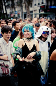 A lively crowd of people walking outdoors; a person with bright blue hair, black crop top, and various necklaces smiles at the center. Next to them, another person wears a white animal-themed hoodie with sunglasses. Background shows a mix of diverse, joyful faces.