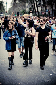 A group of people walking down a street during a crowded event. One person on the left wears a blue denim jacket and black boots, a person in the middle wears all black, holding up a phone, and another on the right also dressed in black. Trees line the street.