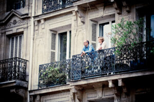 Two people stand on a wrought-iron balcony of a classic stone building, looking out. The man and woman are engaged in conversation, surrounded by greenery. The facade features tall windows with shutters and intricate architectural details.