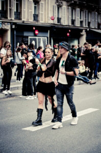 A woman and a man walk along a city street. The woman wears a black outfit with a short skirt and boots, while the man wears a hat, sunglasses, and casual clothes. They are surrounded by onlookers standing on the sidewalk. The background shows buildings and windows.